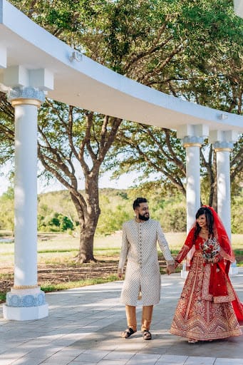 Bride in red and groom in ivory holding hands under a white pergola
