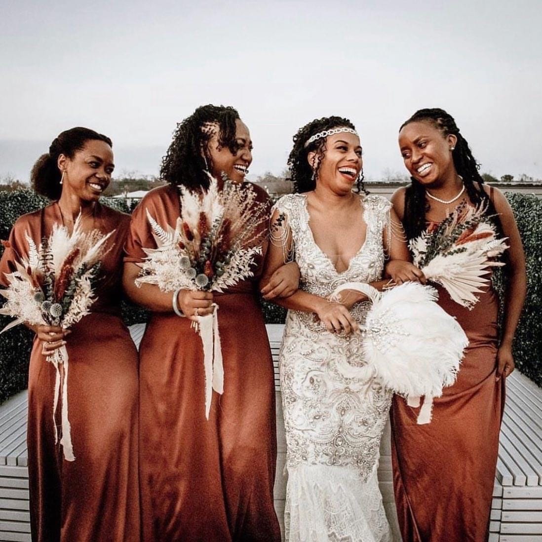 Bride laughing with bridesmaids in rust satin dresses holding pampas bouquets