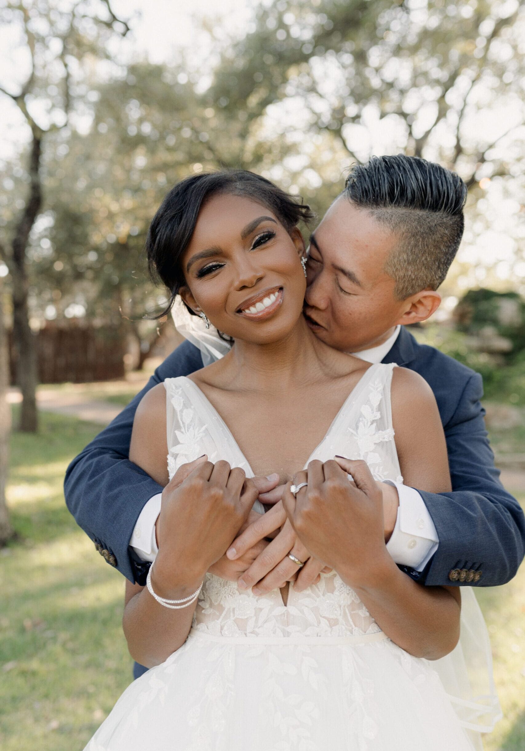 Groom embracing his smiling bride from behind under oak trees