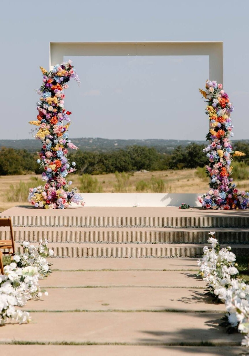 Outdoor ceremony site with two tall floral columns on stone steps and white flowers lining the aisle