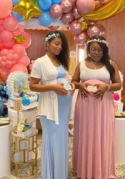 vachon Two expectant mothers in blue and pink dresses and flower crowns in front of a balloon arch